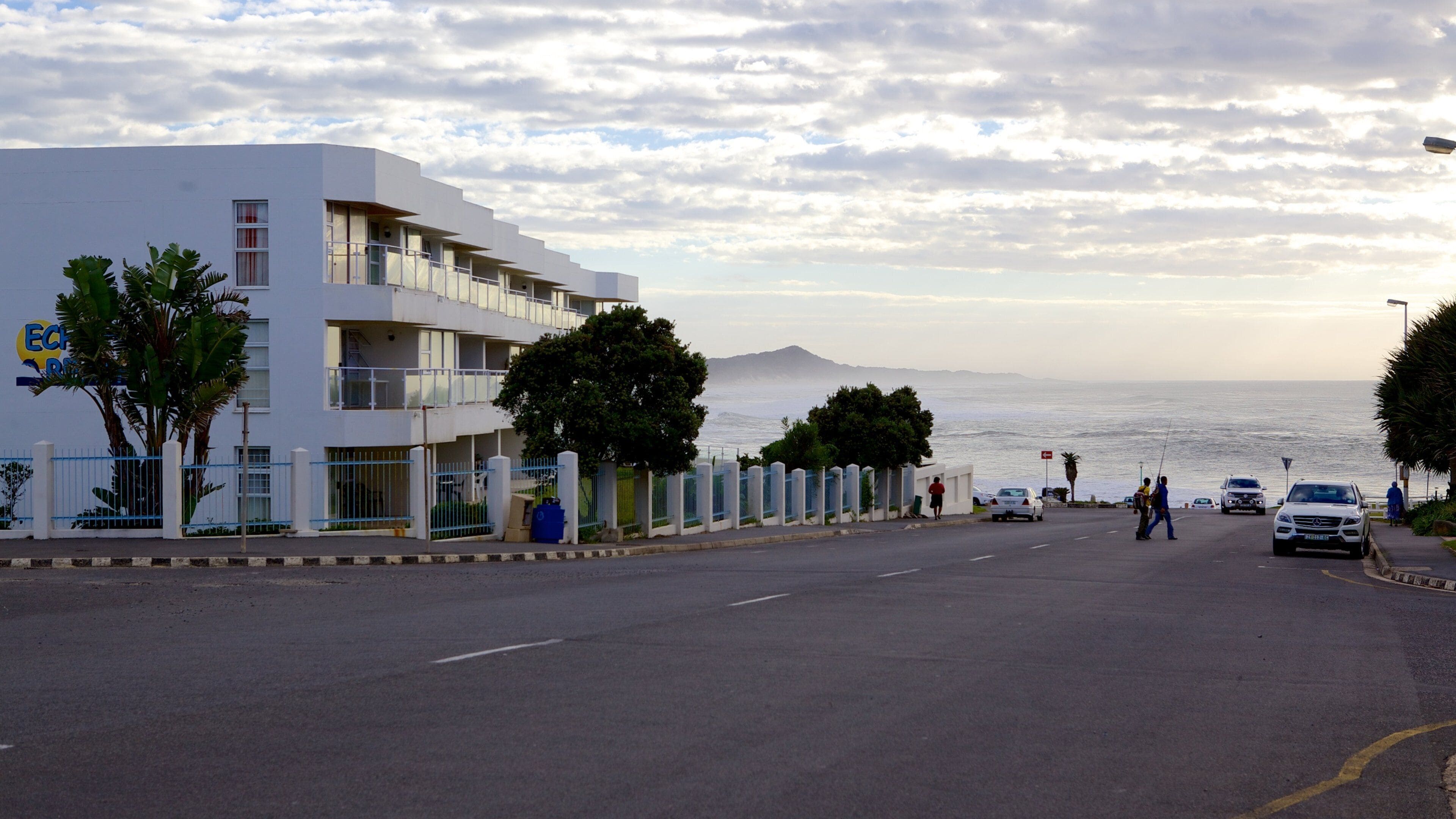 A coastal road with a white building, palm trees, and a view of the sea.