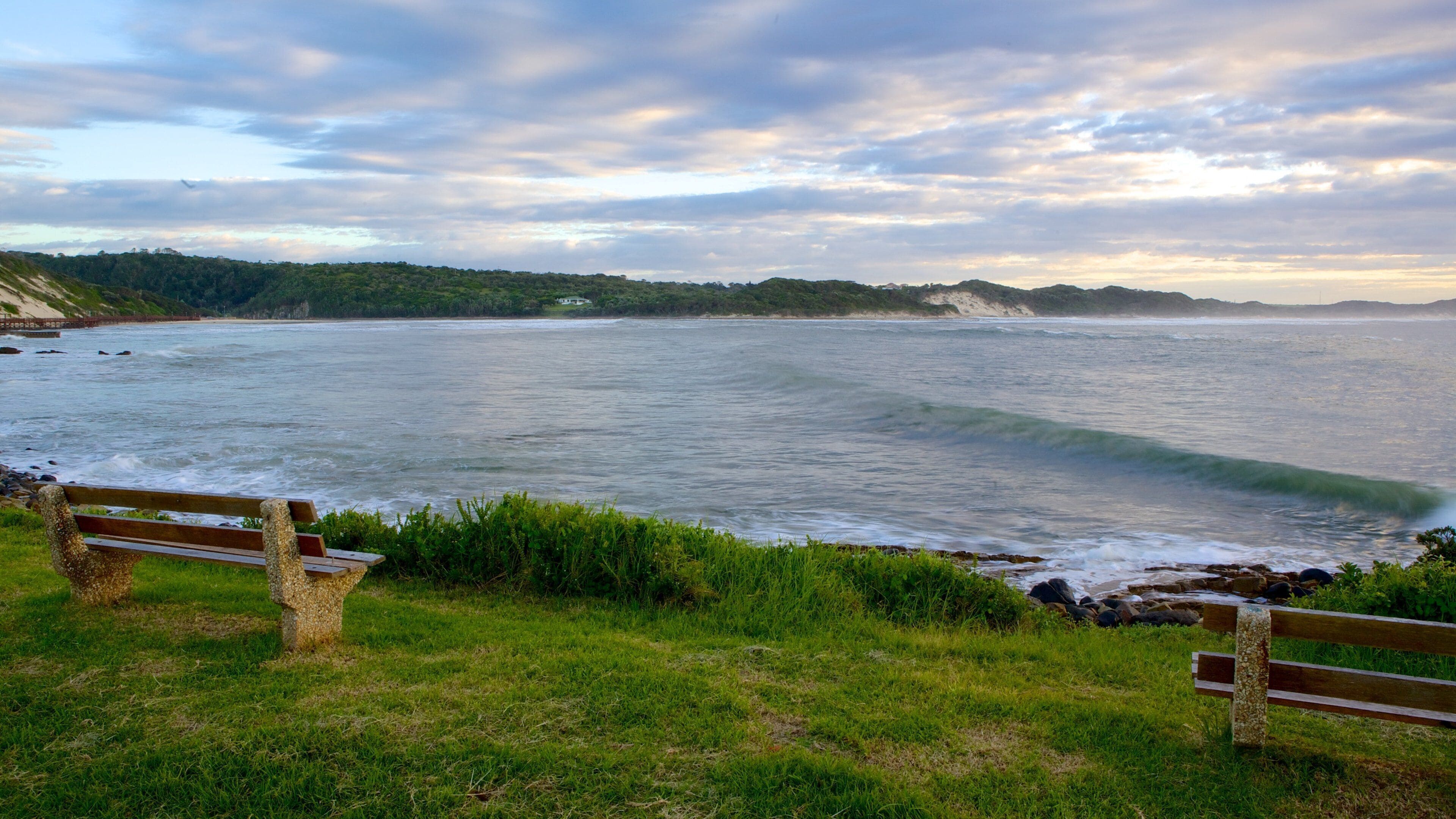 A coastal landscape with a bench overlooking the sea, grassy areas, and distant hills.