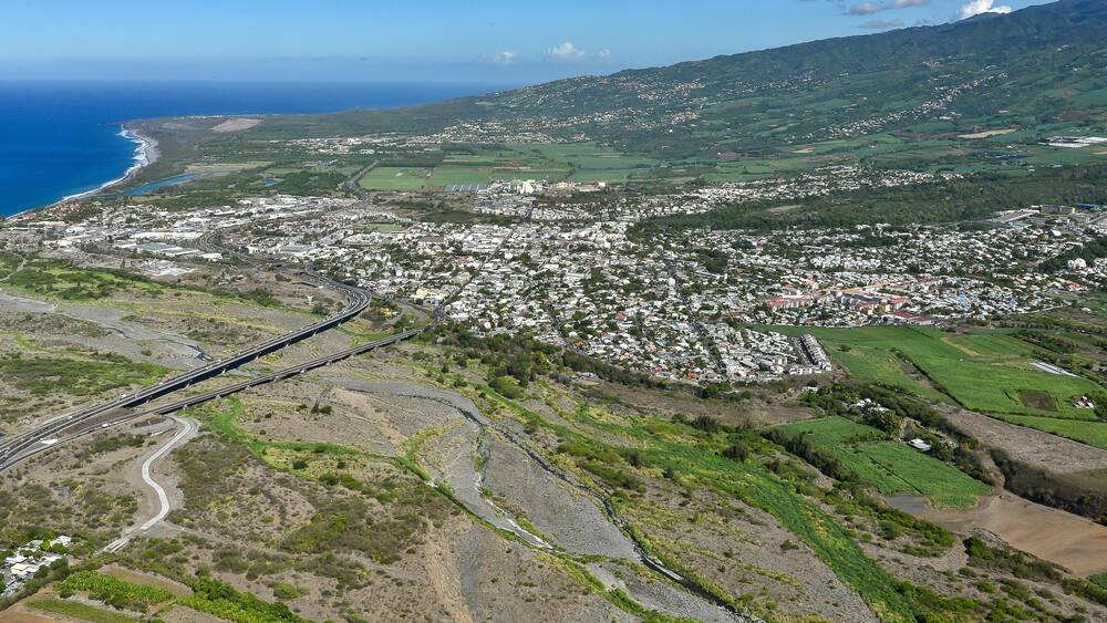 A coastal town with a bridge connecting to the mainland, surrounded by green hills and a clear blue sky.
