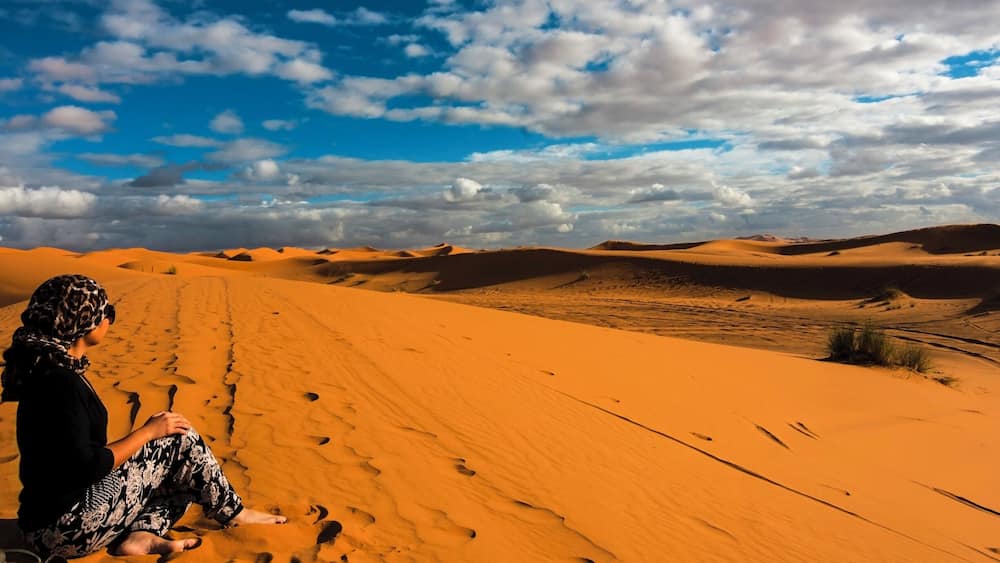 A person sitting on a sand dune with footprints leading towards the horizon.