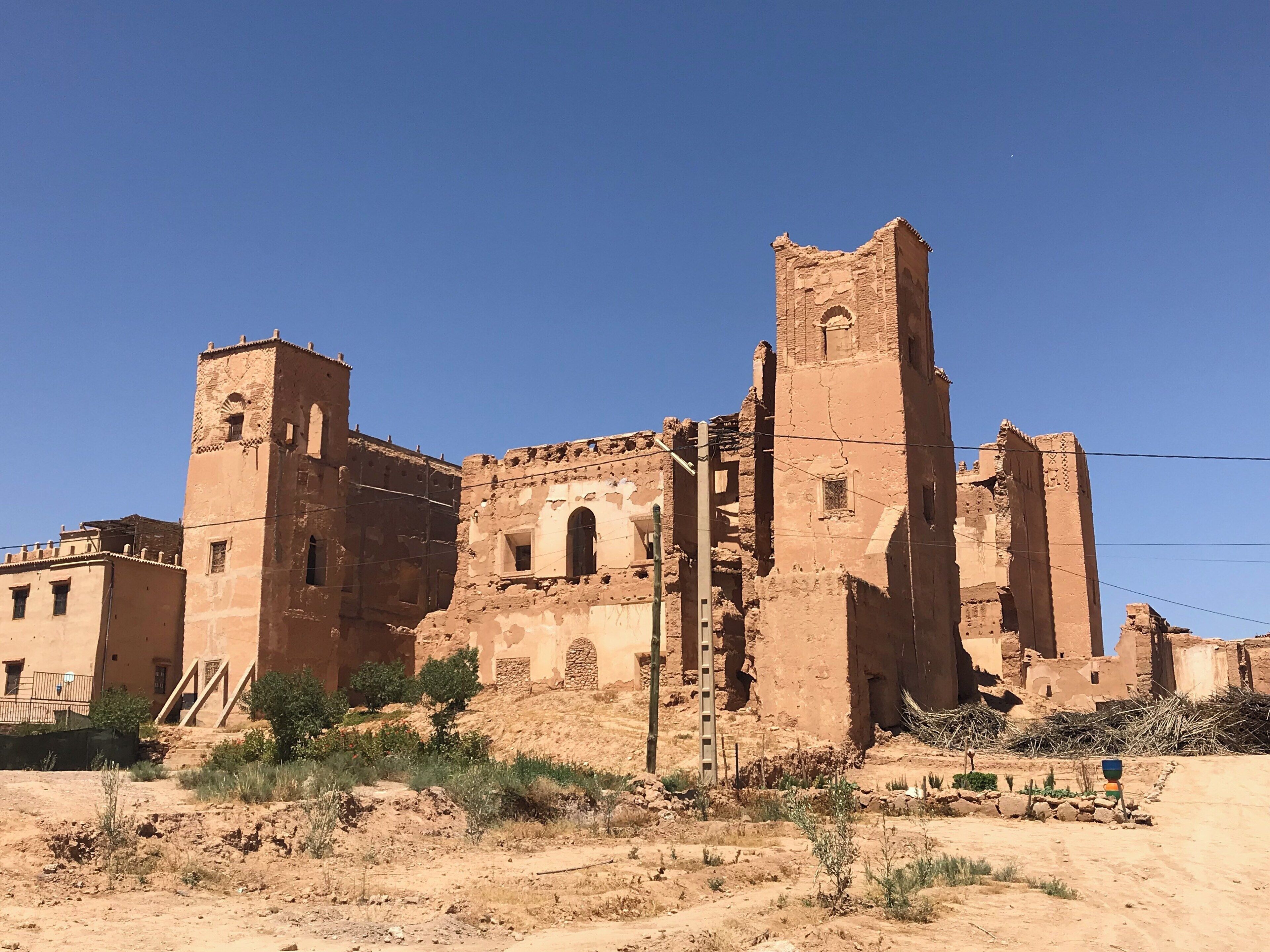 An ancient, partially ruined building with arched windows and a tall tower, surrounded by sparse vegetation and a clear blue sky.