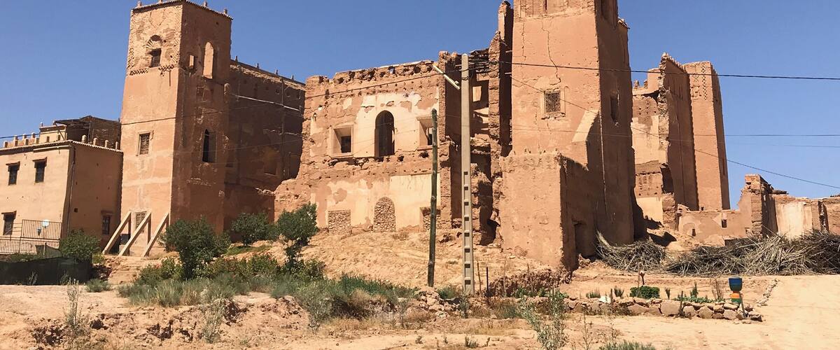 An ancient, partially ruined building with arched windows and a tall tower, surrounded by sparse vegetation and a clear blue sky.