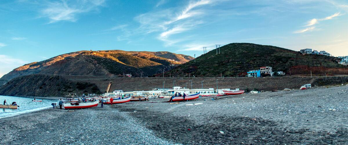 A rocky beach with several boats, a hill with buildings, and a clear sky.