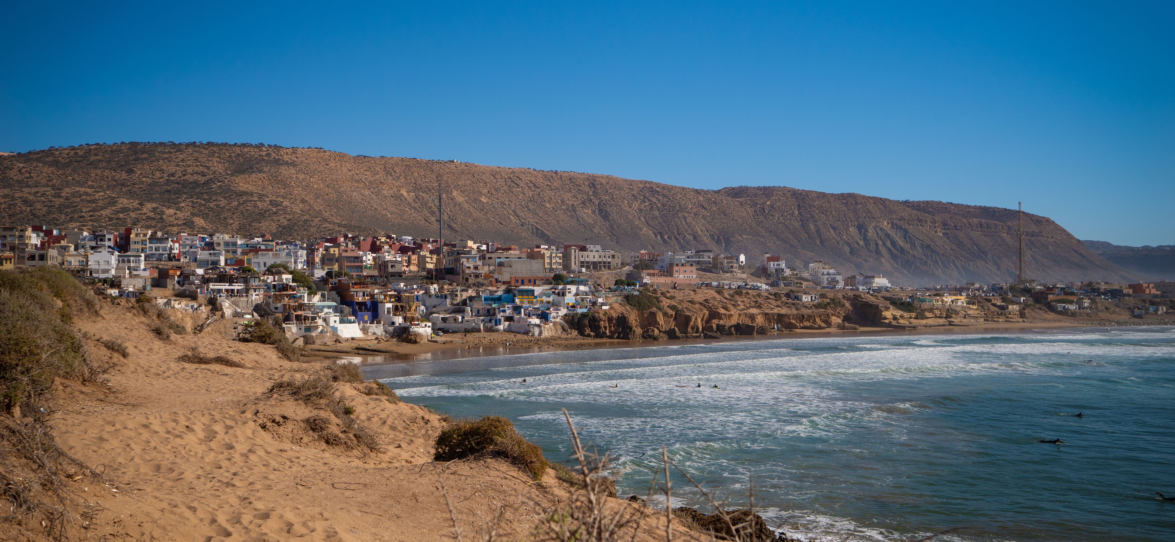 Surfers in water, paddling and waiting for waves, with a charming coastal town of Imsouane and arid hills in the background. Scenic view of colorful buildings overlooking popular Moroccan surf break.