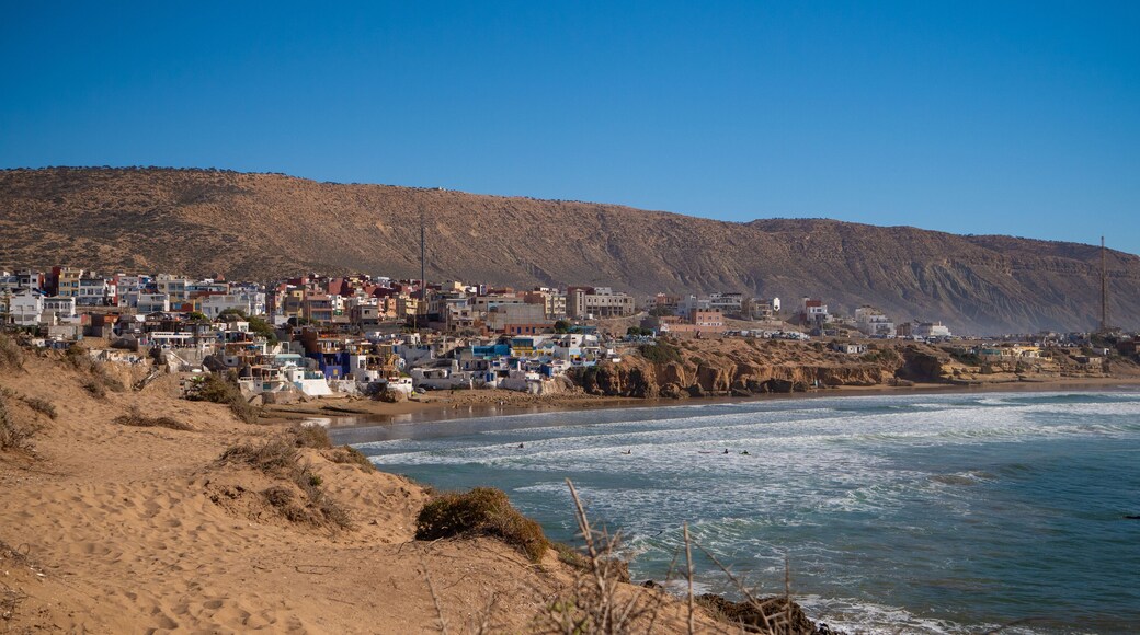 Surfers in water, paddling and waiting for waves, with a charming coastal town of Imsouane and arid hills in the background. Scenic view of colorful buildings overlooking popular Moroccan surf break.
