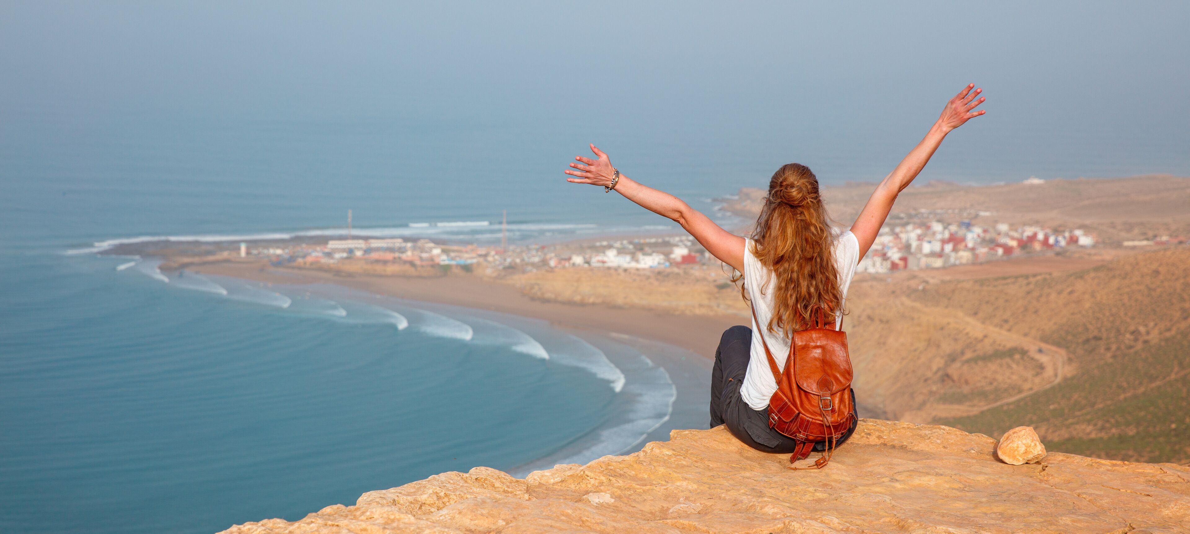A person sitting on a cliff overlooking a coastal town with waves crashing onto the shore.