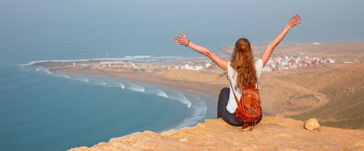 A person sitting on a cliff overlooking a coastal town with waves crashing onto the shore.