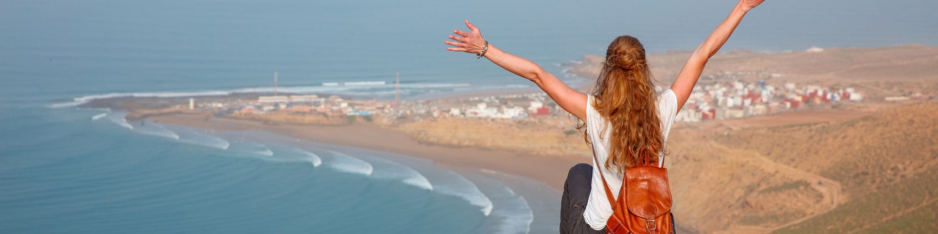 A person sitting on a cliff overlooking a coastal town with waves crashing onto the shore.
