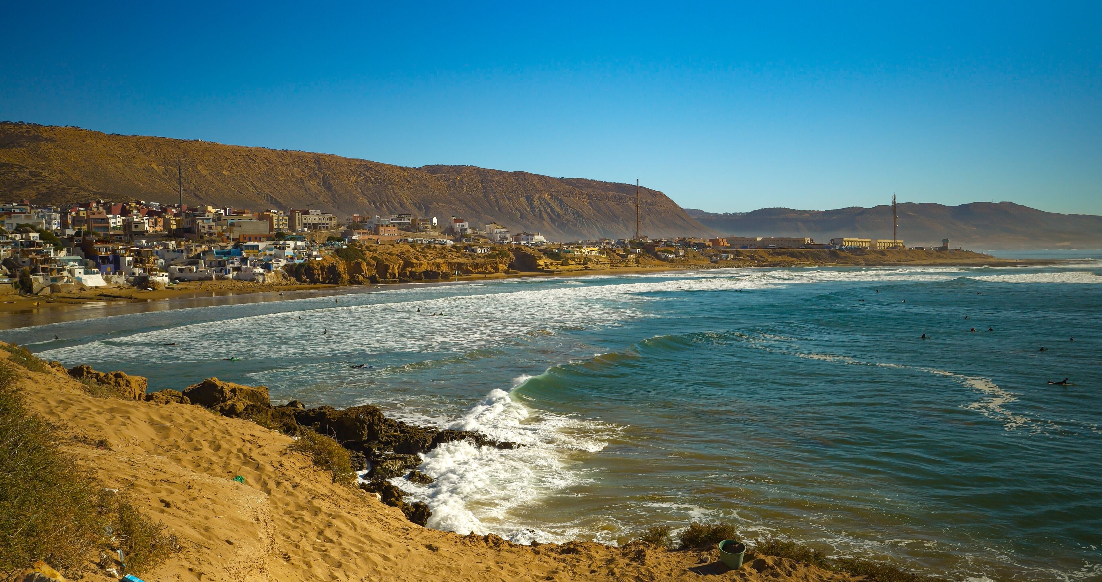 Surfers in the water, paddling and waiting for waves, with a coastal town of Imsouane and arid hills in the background. Peaceful and recreational feeling with a view of the famous Moroccan surf break.