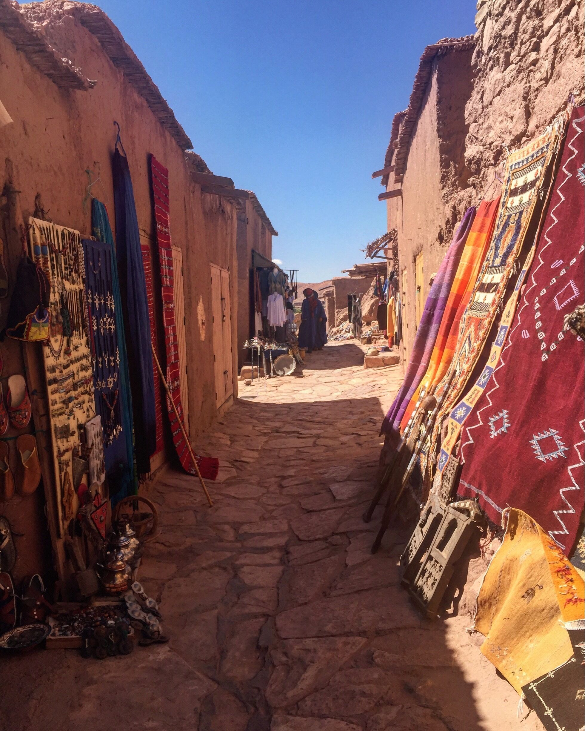 A narrow street with traditional buildings and colorful textiles displayed outside.