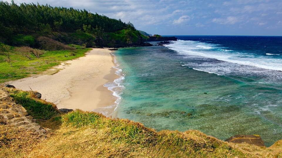 A coastal landscape with a sandy beach, clear turquoise waters, and a forested hillside.