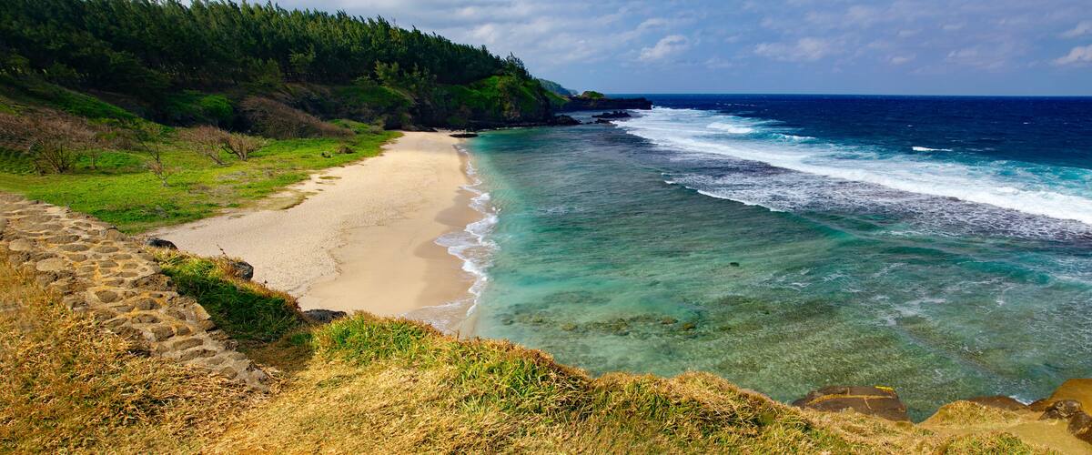 A coastal landscape with a sandy beach, clear turquoise waters, and a forested hillside.