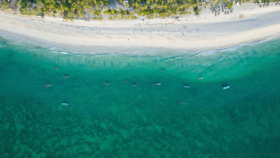 An aerial view of a beach with clear turquoise waters and several small boats anchored near the shore.