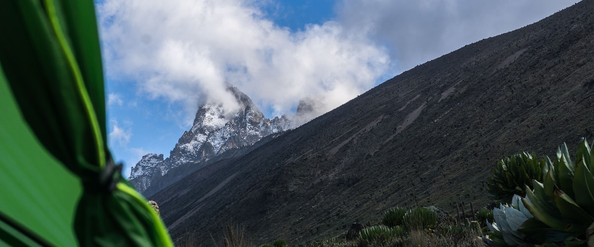 A mountain with snow-capped peaks, a clear blue sky, and some clouds.