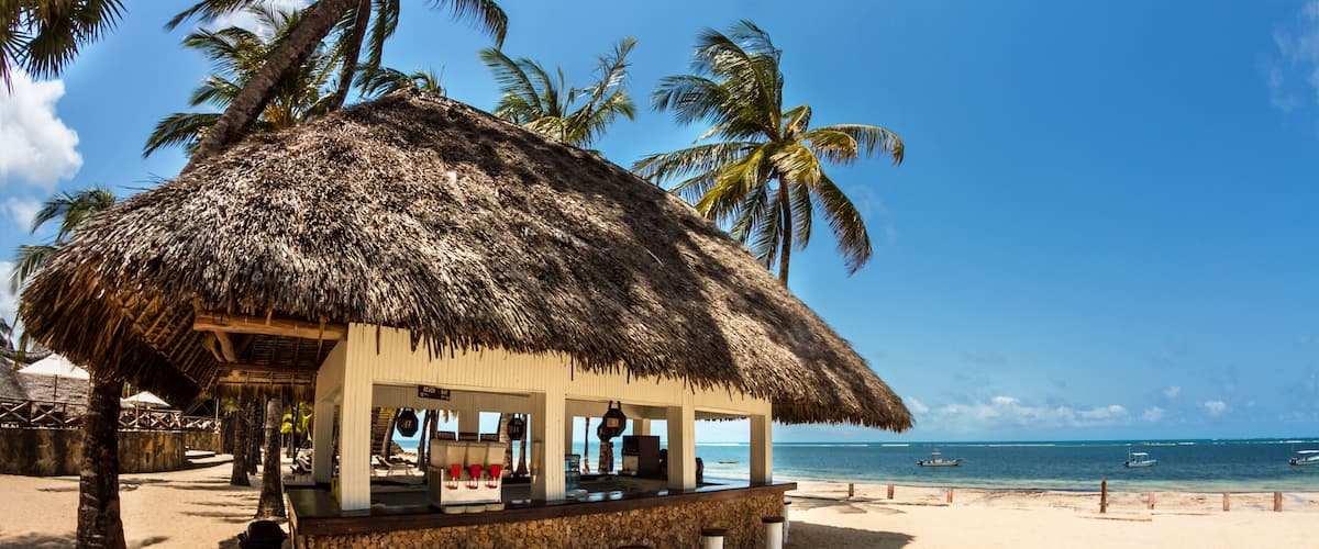 A beach bar with a thatched roof, palm trees, and the ocean in the background.