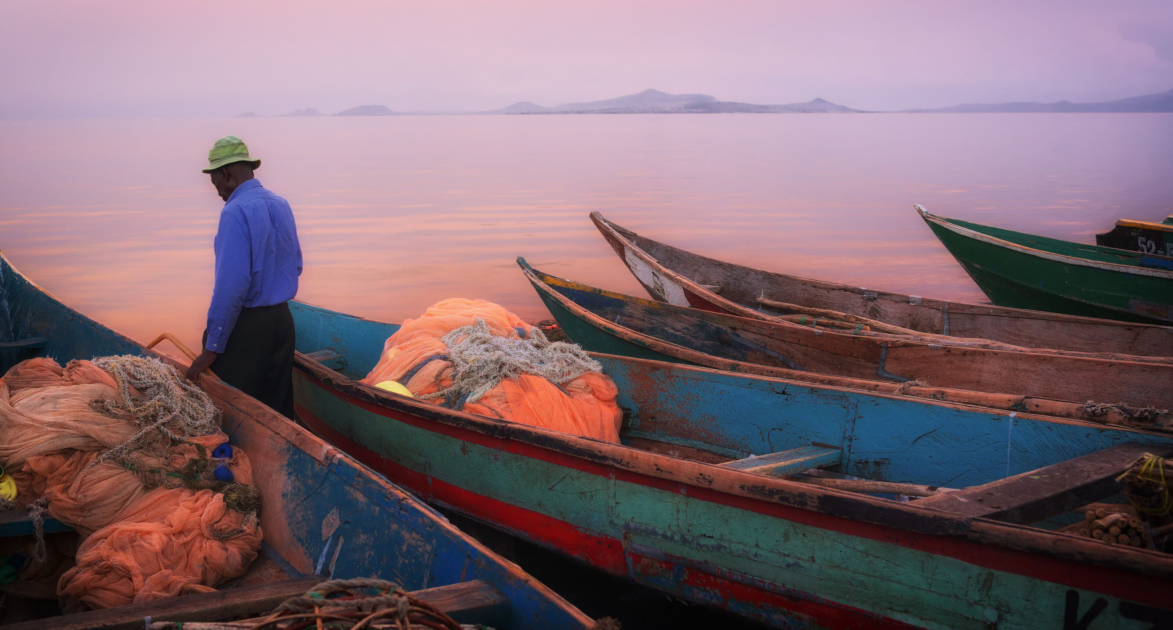 A man in a blue shirt and hat standing by boats at dusk.