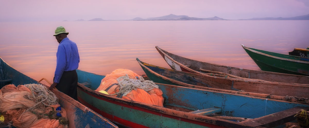 A man in a blue shirt and hat standing by boats at dusk.