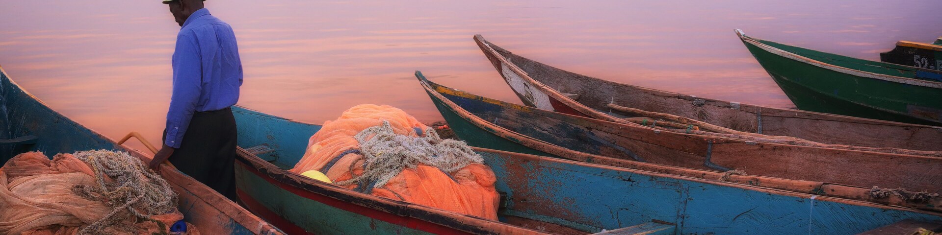 A man in a blue shirt and hat standing by boats at dusk.