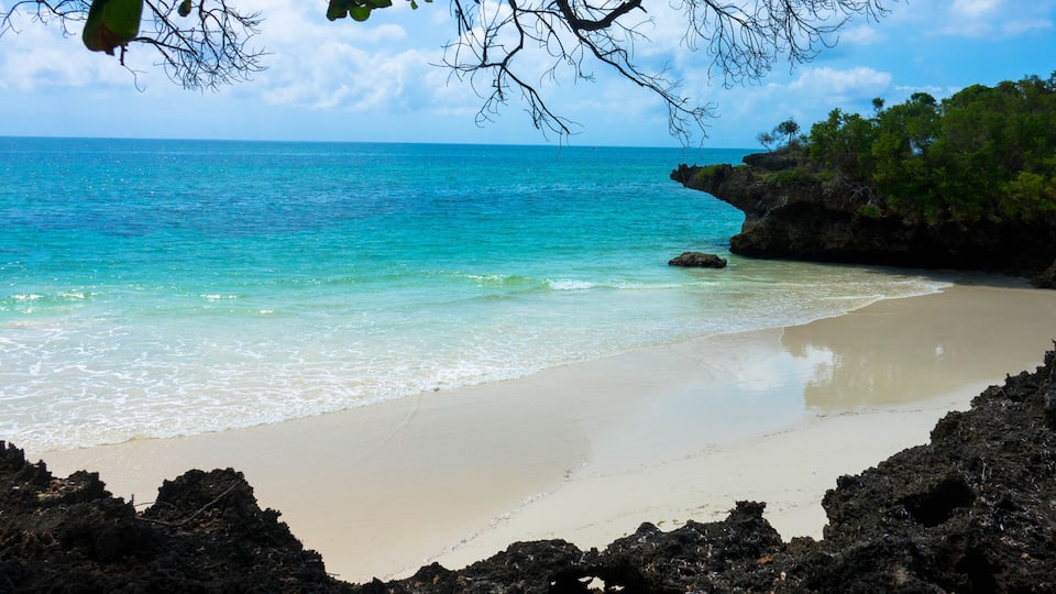 A beach with clear turquoise water, a sandy shore, and rocky outcrops.