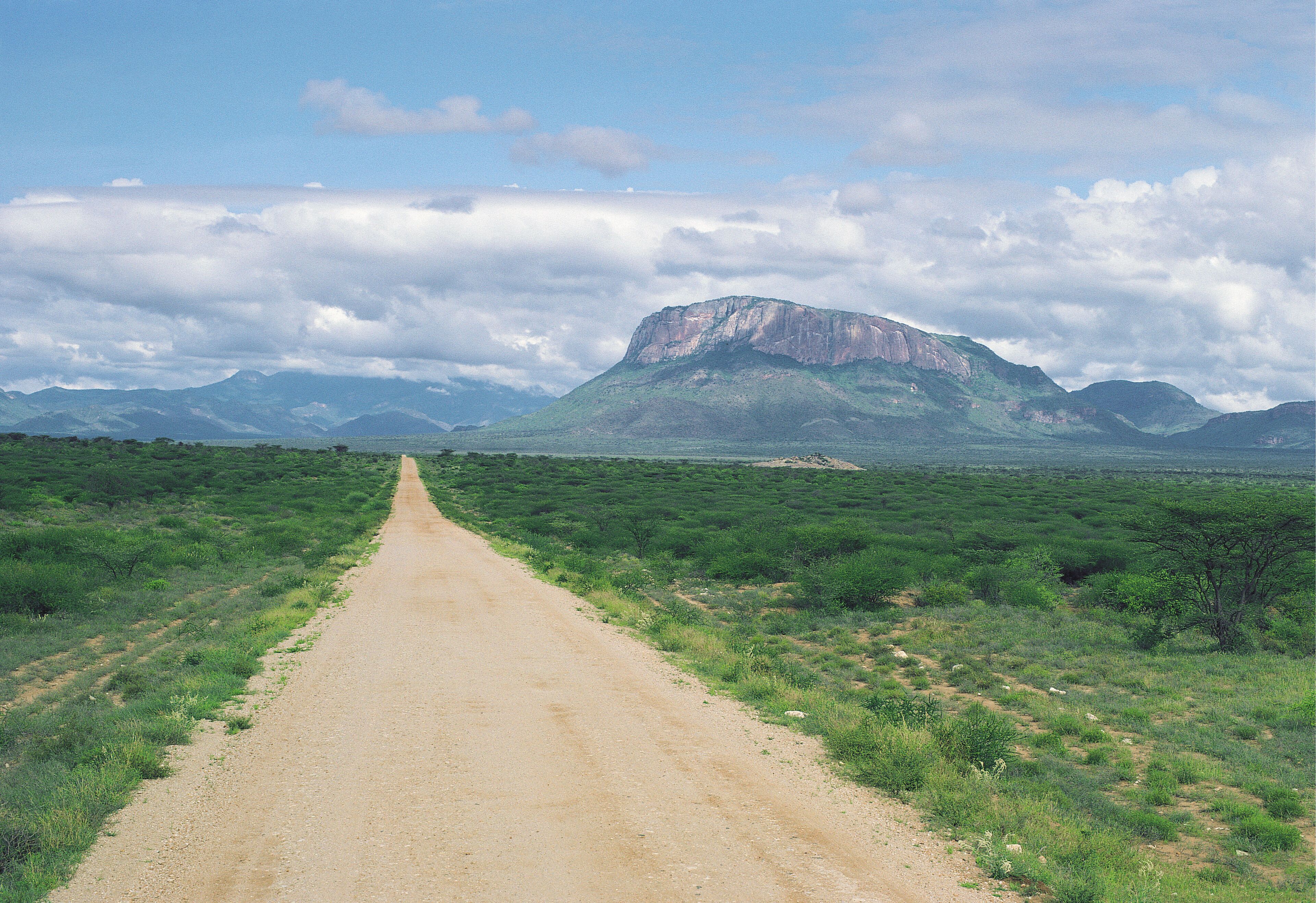 A dirt road leading to a mountain with a flat top, surrounded by green vegetation and under a partly cloudy sky.