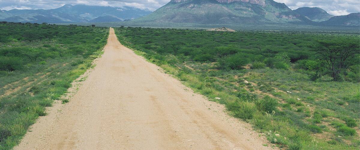 A dirt road leading to a mountain with a flat top, surrounded by green vegetation and under a partly cloudy sky.