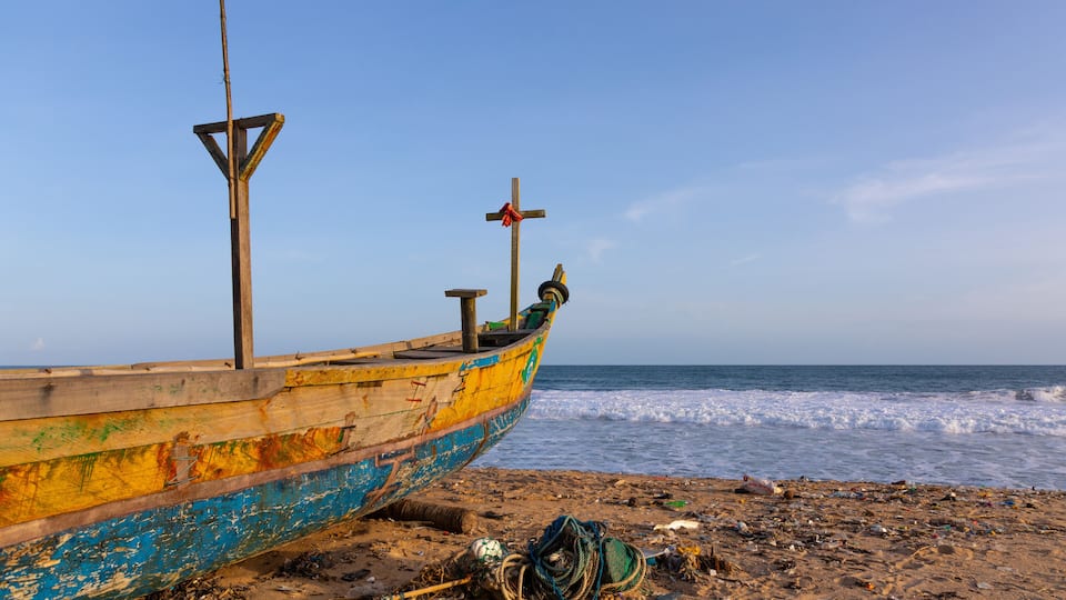 A weathered boat with a cross on its bow, resting on a sandy beach with scattered debris and the ocean in the background.