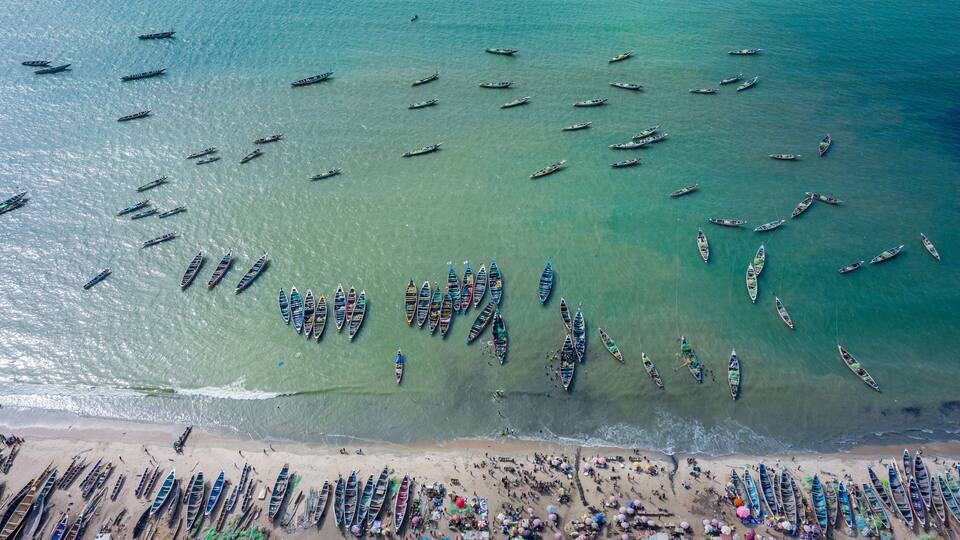 A beach with numerous boats lined up along the shore and a large group of people gathered in the water.