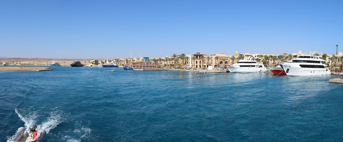 A marina with several yachts docked, a clear blue sky, and a desert landscape in the background.