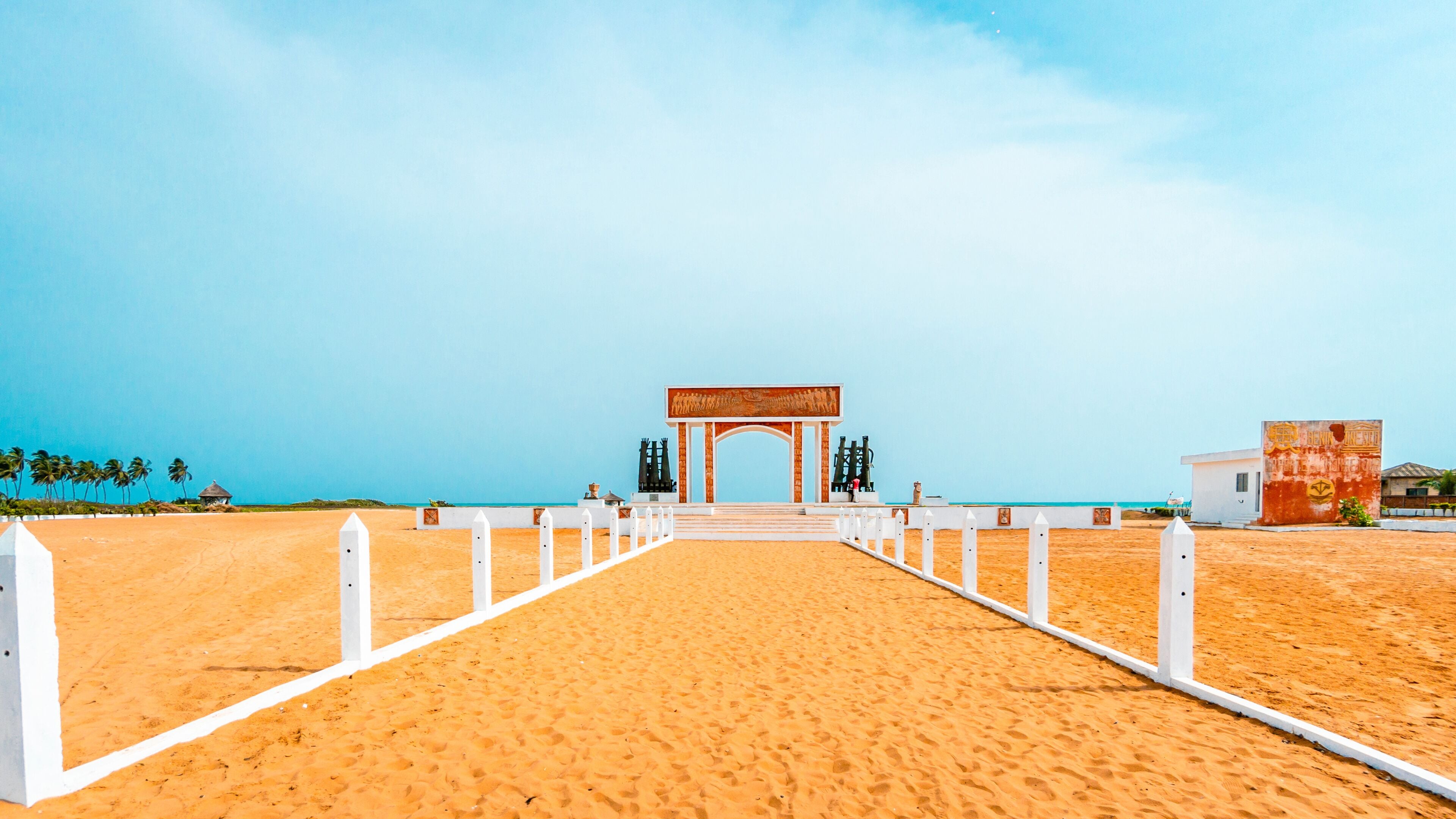 A beach pathway leading to a white archway with a mural, palm trees, and a clear blue sky.