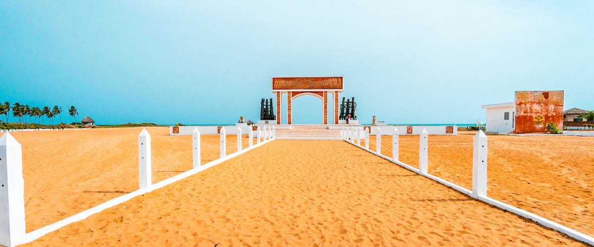 A beach pathway leading to a white archway with a mural, palm trees, and a clear blue sky.