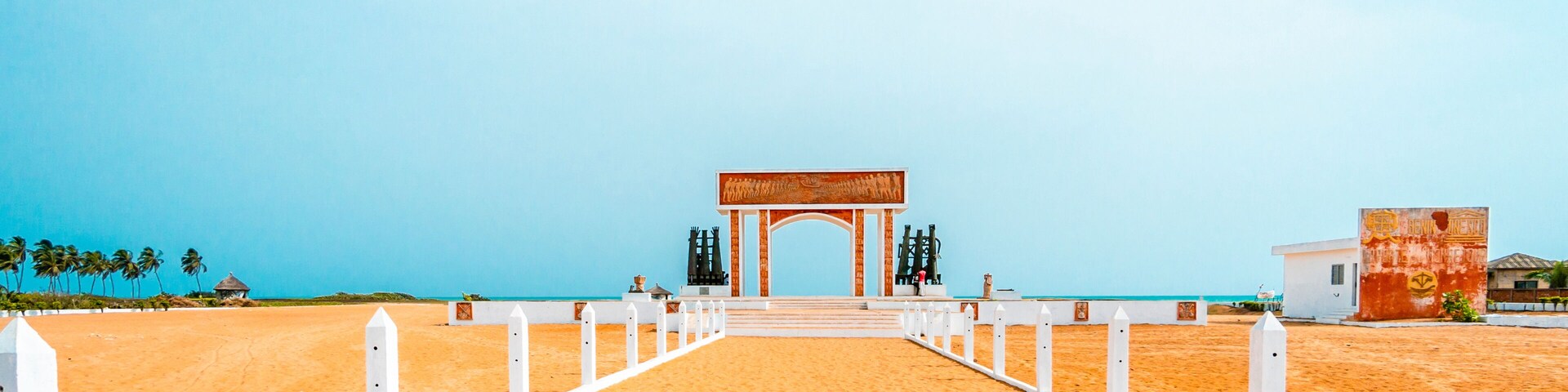 A beach pathway leading to a white archway with a mural, palm trees, and a clear blue sky.