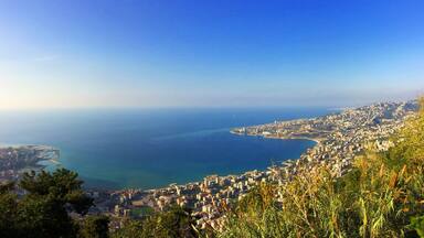 A coastal cityscape with a clear view of the sea, a densely built urban area, and a prominent hill with vegetation.