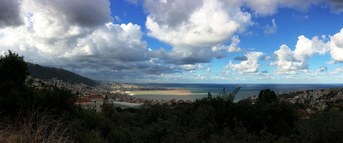 A coastal cityscape with a view of the sea, mountains, and scattered clouds.