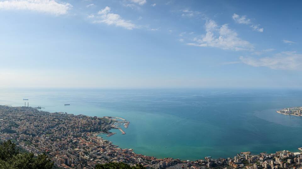 A coastal cityscape with a prominent church on a hill, a clear blue sea, and a person paragliding.