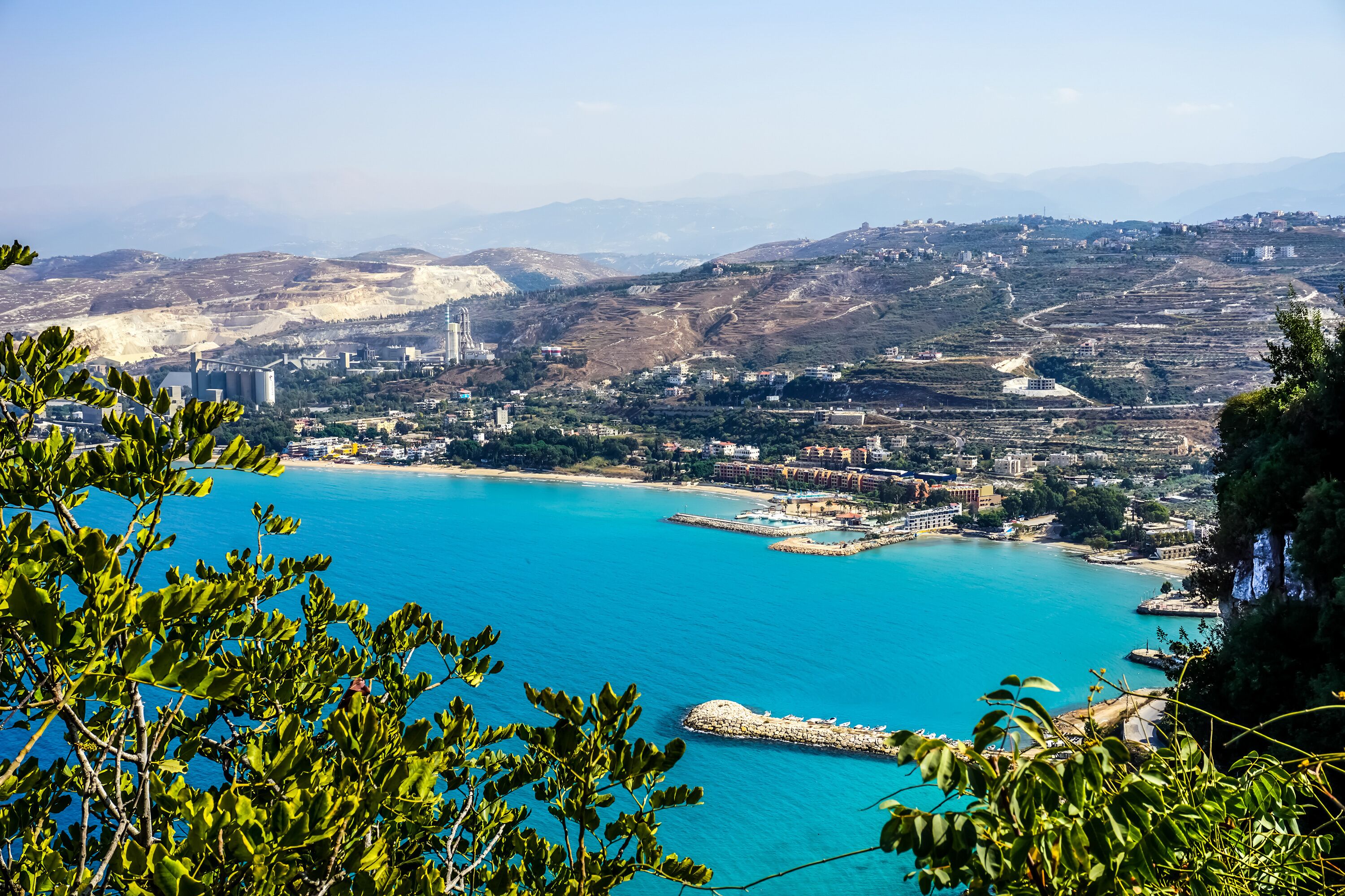 A coastal city with a clear blue sea, green hills, and buildings.