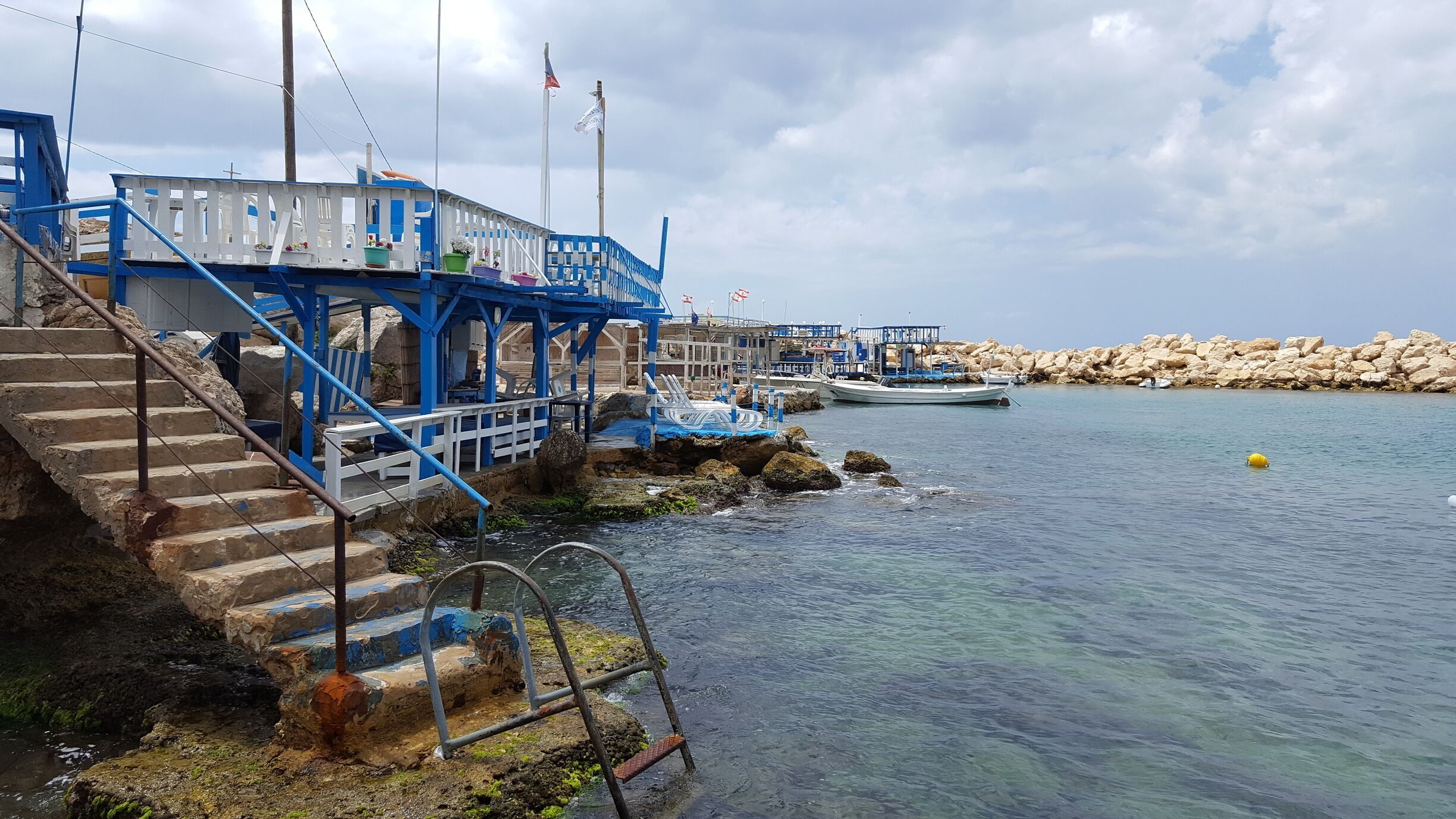 A coastal pier with blue railings, a staircase leading down to the water, and a rocky shoreline.
