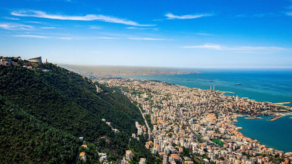 A coastal cityscape with a dense urban area, a prominent hill with greenery, and a clear blue sky.