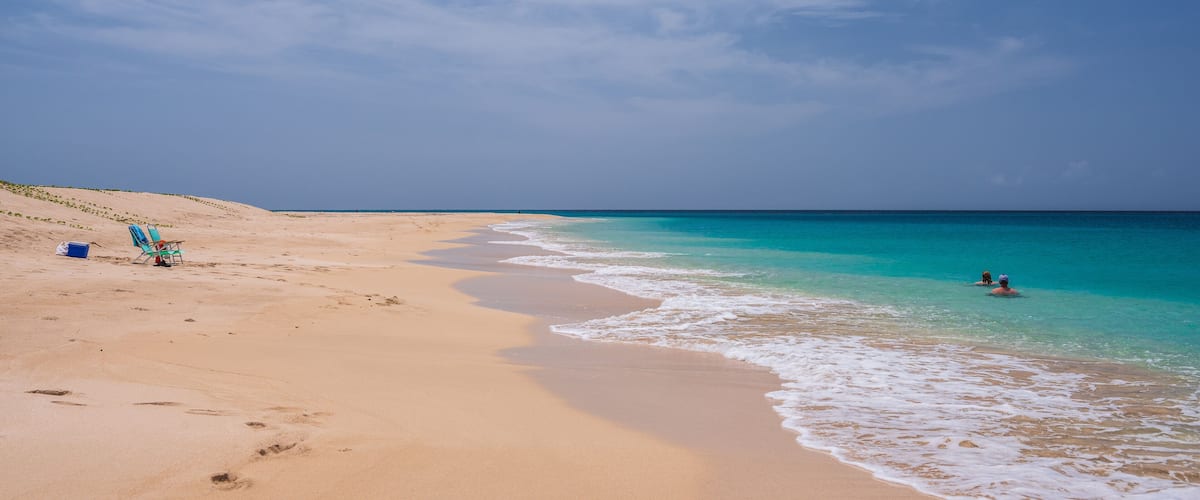 A sandy beach with turquoise water, a clear sky, and footprints leading to a blue chair and a cooler.