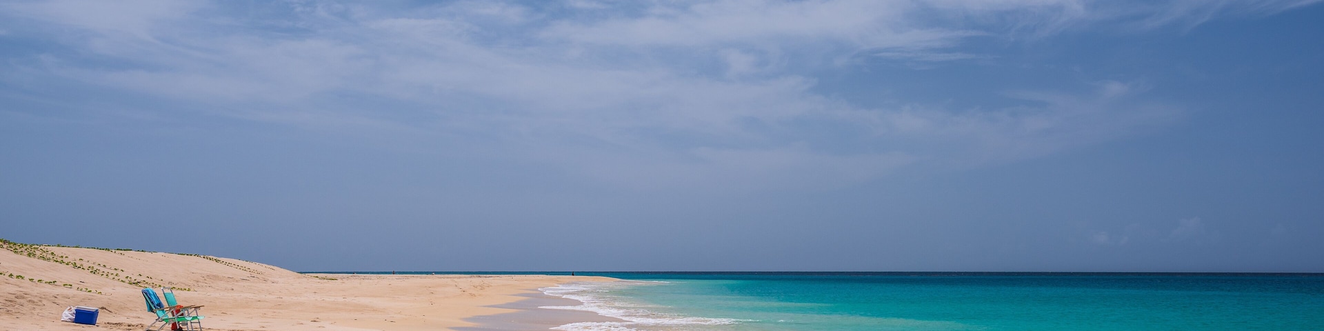 A sandy beach with turquoise water, a clear sky, and footprints leading to a blue chair and a cooler.