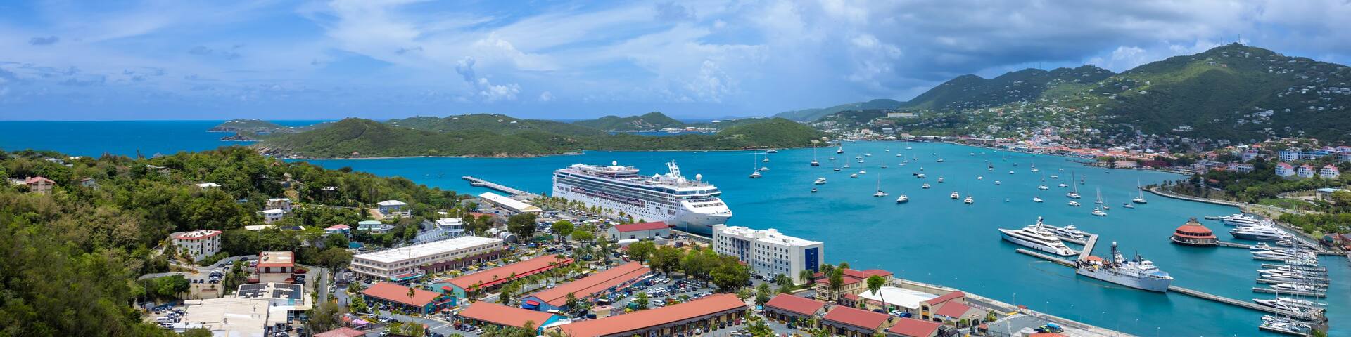 A coastal town with a cruise ship docked, a marina with boats, and a mountainous backdrop.