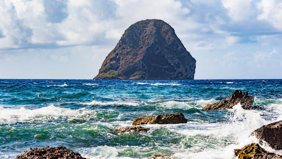 A rocky coastline with waves crashing against the rocks and a prominent mountain in the distance.