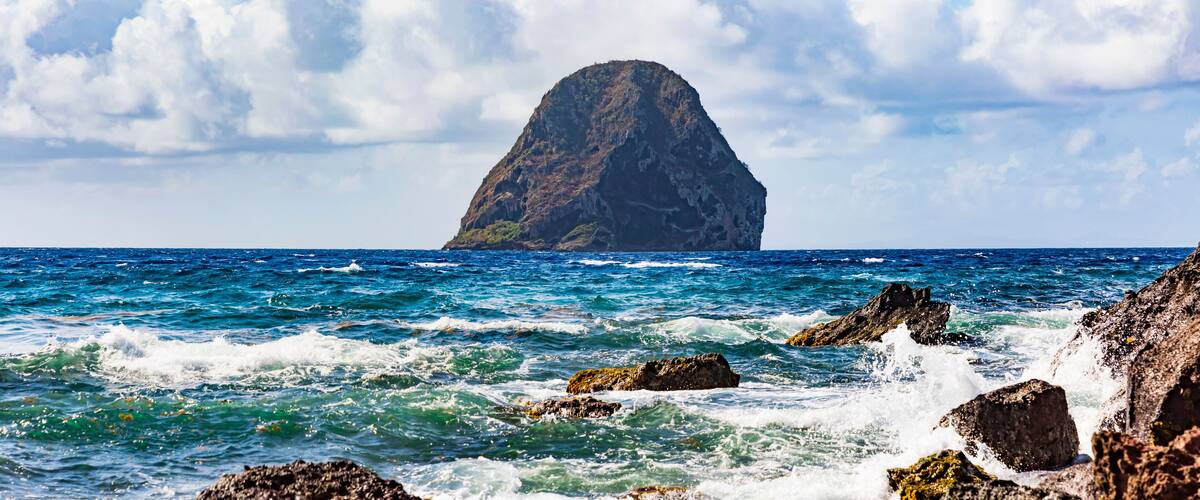 A rocky coastline with waves crashing against the rocks and a prominent mountain in the distance.