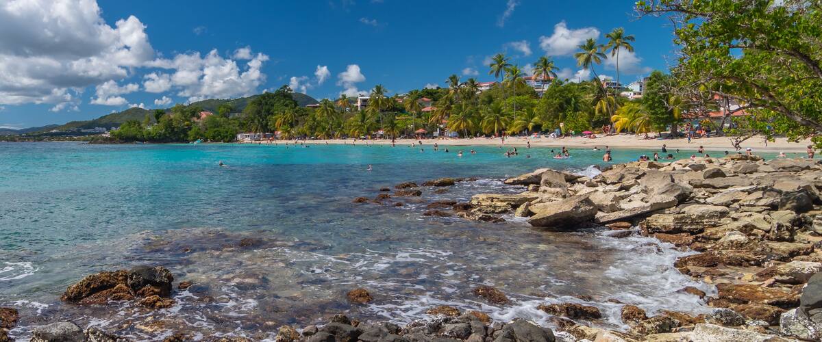 A beach with clear turquoise water, rocky shoreline, palm trees, and buildings in the background.