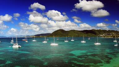 A coastal scene with sailboats anchored in clear turquoise waters, surrounded by lush green hills and a clear blue sky with scattered clouds.