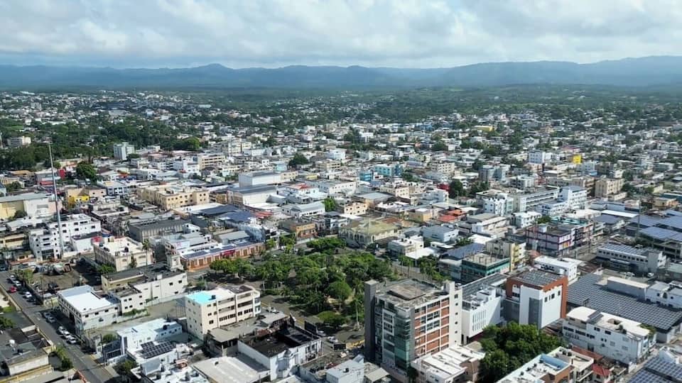 A densely populated urban area with a mix of residential and commercial buildings, surrounded by greenery and distant mountains.