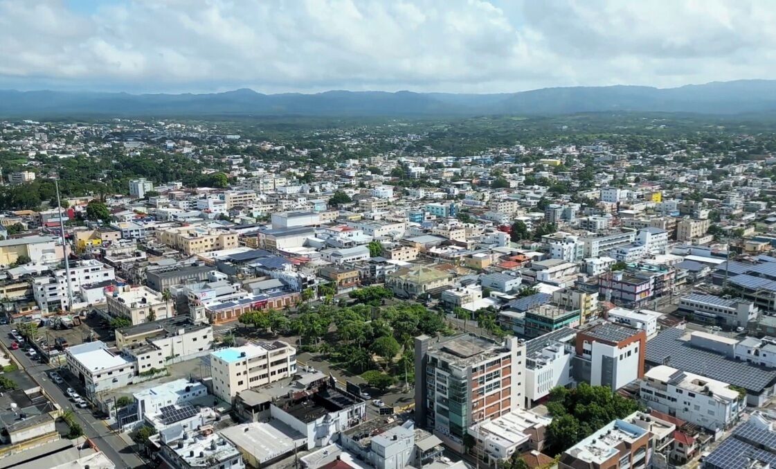 A densely populated urban area with a mix of residential and commercial buildings, surrounded by greenery and distant mountains.