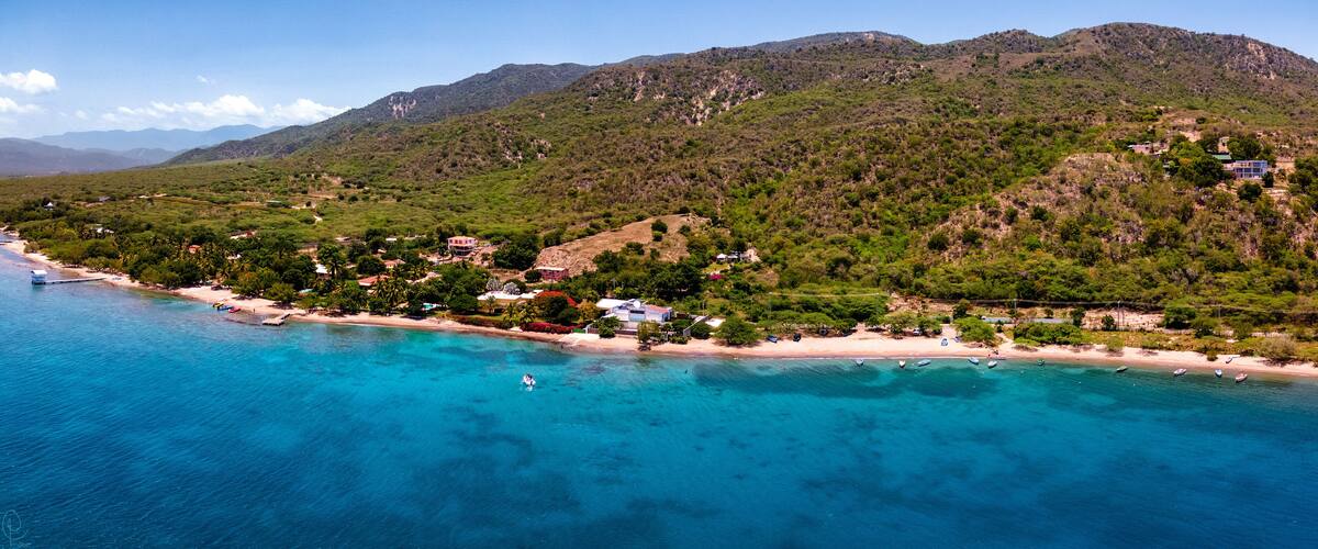 A coastal landscape with a beach, clear blue waters, and a mountainous backdrop.