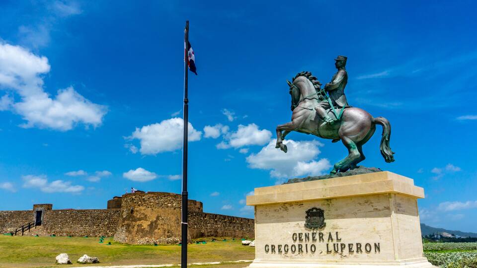 A statue of General Gregorio Luperon on a horse, with a flagpole and a stone wall in the background.