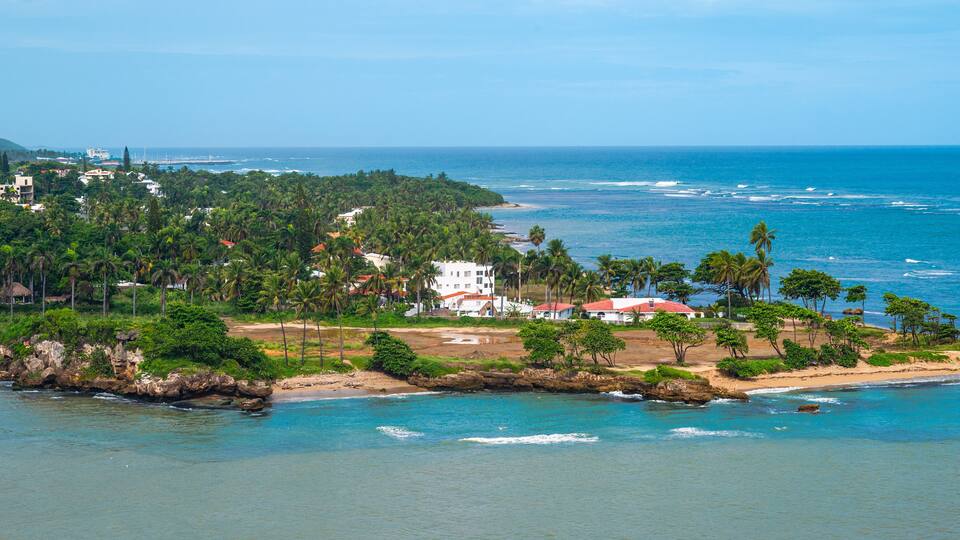 A coastal landscape with a beach, palm trees, and buildings.