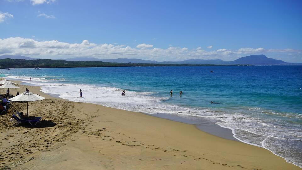 A beach with clear blue water, sandy shore, and distant mountains.